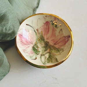 Puede incluir: Un pequeño cuenco redondo de cerámica con un diseño floral. El cuenco presenta un interior color crema con flores rosas y hojas verdes. El borde está adornado con oro y el exterior tiene una banda verde. Probablemente se utiliza para servir pequeñas porciones de comida o como objeto decorativo.