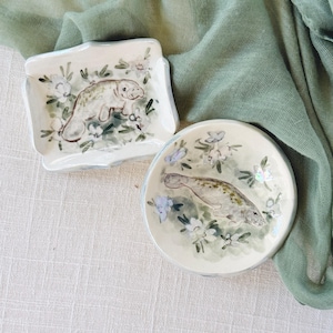 Peut inclure: Deux plats en céramique avec des illustrations de lamantins. L'un est carré avec des bords festonnés, l'autre est rond. Les deux ont une base blanche avec des motifs floraux et de lamantins aux couleurs douces. Les plats sont sur une surface claire avec un tissu vert.