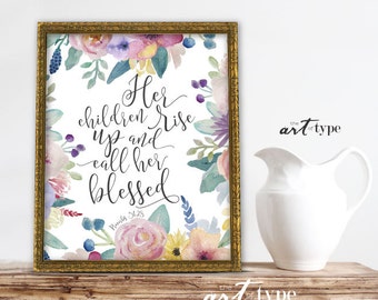 Pode incluir: Uma impressão de arte emoldurada com uma moldura dourada. A impressão apresenta arranjos florais em aquarela em cores pastel em torno do texto "Her children rise up and call her blessed". A arte é exibida em uma superfície de madeira.