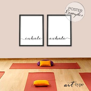 May include: Two black-framed posters with the words "inhale" and "exhale" in cursive script. The posters are white with black text and are displayed on a pale pink wall. Yoga mats and bolsters are in the foreground.