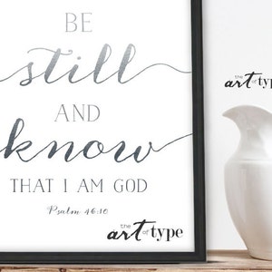 K&ouml;nnte beinhalten: Ein wei&szlig;er druckbarer Kunstdruck mit silbernem Metallic-Design. Der Druck zeigt den Text "Be still and know that I am God" mit dem Schriftzitat "Psalm 46:10". Der Druck ist mit einem schwarzen Rahmen gerahmt.