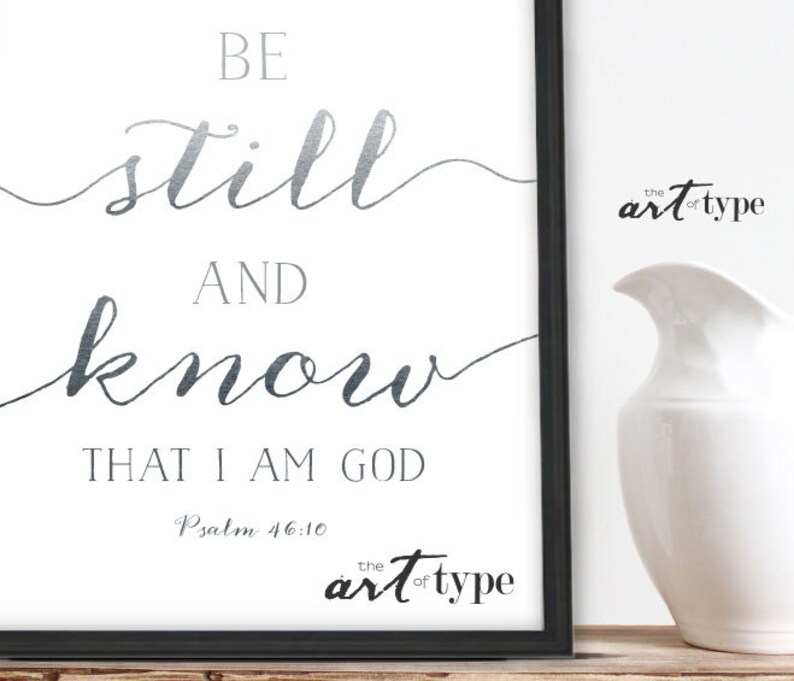 K&ouml;nnte beinhalten: Ein wei&szlig;er druckbarer Kunstdruck mit silbernem Metallic-Design. Der Druck zeigt den Text "Be still and know that I am God" mit dem Schriftzitat "Psalm 46:10". Der Druck ist mit einem schwarzen Rahmen gerahmt.