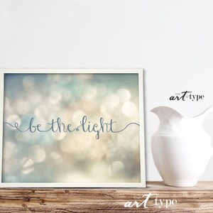 May include: A framed print with a blurred background of white, blue, and green lights. The text "be the light" is written in a cursive font in blue.