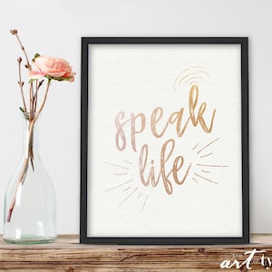 May include: A framed print with a white background and black frame. The print features the text "speak life" in gold script lettering with a sunburst design behind the text.