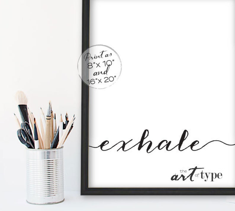 May include: A black framed print with the word "exhale" in elegant script. The phrase "the art of type" is below. A circular graphic indicates the print sizes are 20cm x 25cm and 40cm x 50cm. A metal container holds art supplies.