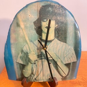 May include: An arched wall clock featuring a printed image of a person in a pinstriped baseball uniform holding a bat. The clock face has a blue resin border and three black hands at the center. It is supported by a small brown display stand.