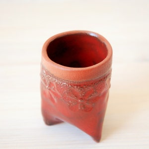 May include: A small, red, ceramic planter with a decorative design around the rim. The planter has three legs and is ideal for small plants.