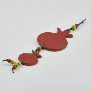 Puede incluir: Un adorno colgante decorativo con dos formas de granada de color marr&oacute;n rojizo. Las granadas est&aacute;n conectadas por una cuerda verde, adornadas con cuentas de madera rojas, amarillas y verdes. El adorno est&aacute; sobre un fondo blanco.