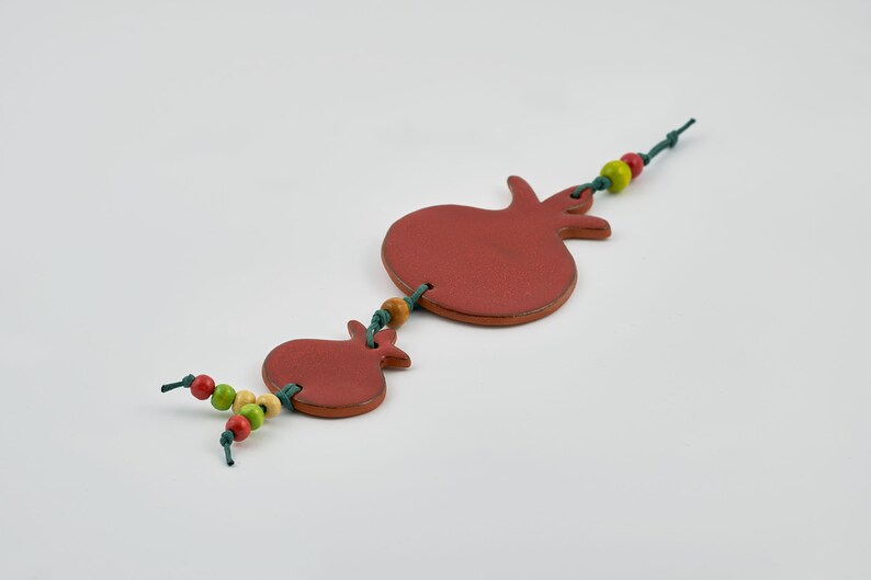 Puede incluir: Un adorno colgante decorativo con dos formas de granada de color marr&oacute;n rojizo. Las granadas est&aacute;n conectadas por una cuerda verde, adornadas con cuentas de madera rojas, amarillas y verdes. El adorno est&aacute; sobre un fondo blanco.