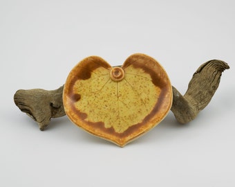 May include: A heart-shaped ceramic dish with a leaf design in shades of yellow and brown. The dish has a small, round indentation in the center.