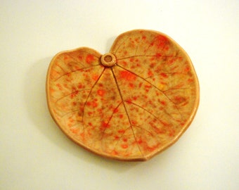 May include: A ceramic leaf-shaped dish with a light brown glaze and orange and yellow speckles. The dish has a small hole in the center.