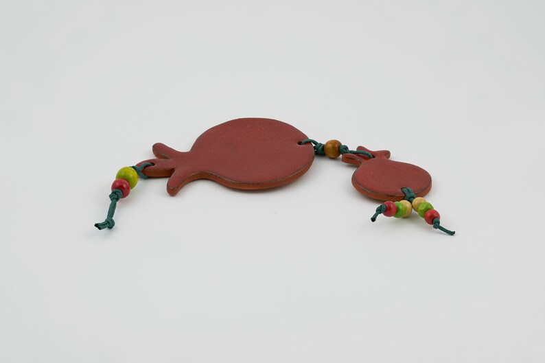 Puede incluir: Adorno en forma de granada de color terracota con una cuerda de cuentas de colores y cuerda verde. El adorno tiene un aspecto r&uacute;stico y hecho a mano, probablemente para decoraci&oacute;n.
