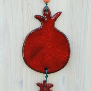 Puede incluir: Adorno artesanal de cer&aacute;mica roja con forma de granada, con superficie texturizada y contorno marr&oacute;n oscuro. El adorno est&aacute; suspendido por una cuerda verde y presenta una forma de granada m&aacute;s peque&ntilde;a debajo. Fondo de madera clara.