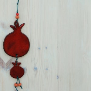 Puede incluir: Dos adornos rojos en forma de granada colgados sobre un fondo de madera clara. El adorno m&aacute;s grande est&aacute; en la parte superior, con uno m&aacute;s peque&ntilde;o debajo, conectados por una cuerda verde y cuentas naranjas. Los adornos tienen un acabado brillante.