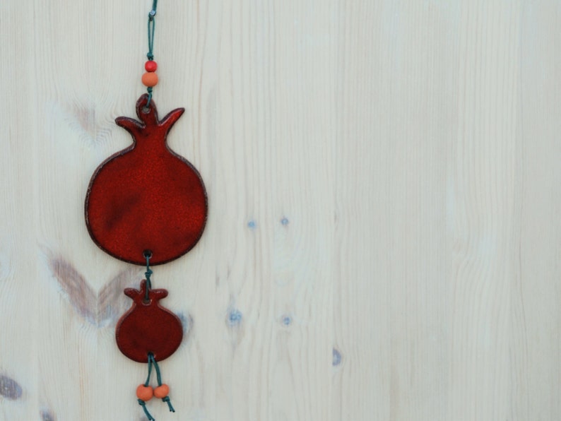 Puede incluir: Dos adornos rojos en forma de granada colgados sobre un fondo de madera clara. El adorno m&aacute;s grande est&aacute; en la parte superior, con uno m&aacute;s peque&ntilde;o debajo, conectados por una cuerda verde y cuentas naranjas. Los adornos tienen un acabado brillante.