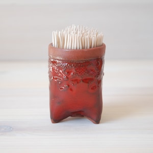 May include: A red ceramic toothpick holder with a textured surface and three legs. The holder is filled with toothpicks.