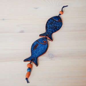 Puede incluir: Dos adornos de cerámica con forma de pez azul conectados por una cuerda, con cuentas naranjas y azules. Los peces tienen una superficie texturizada y un borde naranja. Los adornos se muestran sobre una superficie de madera clara.