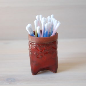 May include: A red ceramic holder with three legs, filled with cotton buds of various colours.