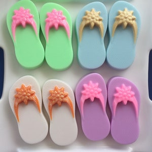 May include: Six pairs of colorful flip-flop shaped soaps with glitter accents. The soaps are arranged in two rows of three on a white tray.