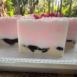Pode incluir: Três sabonetes artesanais retangulares com um design em camadas. A camada superior é branca, transicionando para rosa no meio, e uma base bege claro com inclusões marrons escuras. Os sabonetes são dispostos em uma bandeja branca decorativa.