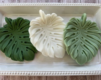 May include: Three decorative monstera leaf-shaped dishes, one white and two green, on a white tray with a beaded edge.