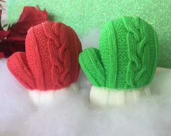 May include: Two knitted mitten bath bombs, one red and one green, sitting on a white fluffy surface.