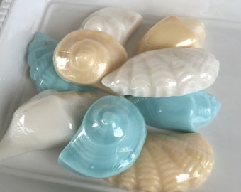 May include: A white ceramic dish with a beaded rim holds a collection of seashell-shaped soaps. The soaps are in shades of white, cream, and light blue.
