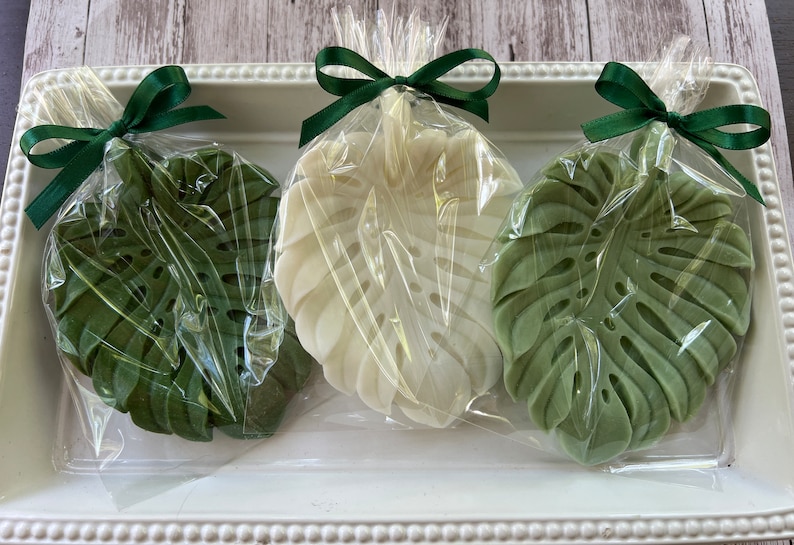 Pode incluir: Tr&ecirc;s sabonetes em forma de folha, verde e branco, embrulhados em pl&aacute;stico transparente com la&ccedil;os de fita verde.