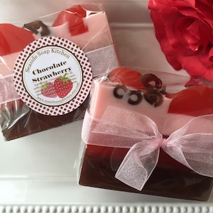 Pode incluir: Duas barras de sabonete artesanal embrulhadas em celofane e amarradas com uma fita rosa. O sabonete é em camadas de tons de marrom, rosa e vermelho, com um formato de coração no topo. O rótulo diz "Seaside Soap Kitchen Chocolate Strawberry".