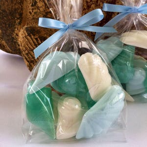 May include: A clear plastic bag tied with a light blue ribbon contains a variety of small, handmade soap bars. The soaps are shaped like seashells and are in shades of light blue and green.