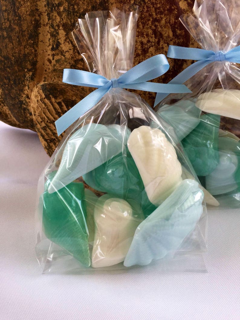 May include: A clear plastic bag tied with a light blue ribbon contains a variety of small, handmade soap bars. The soaps are shaped like seashells and are in shades of light blue and green.