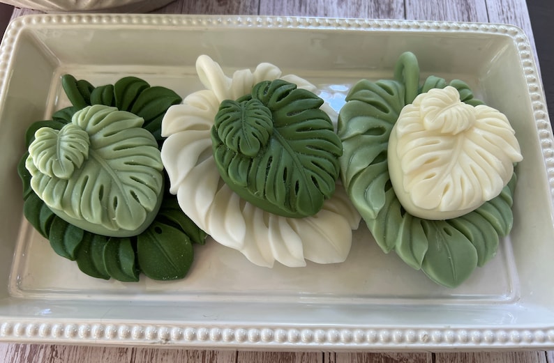 May include: Four green and white soap bars shaped like tropical leaves on a white tray. The soaps are arranged in a row and have a raised design.