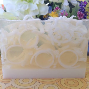 May include: A rectangular bar of soap with a clear, translucent top section filled with white, ring-shaped and shredded coconut pieces. The bottom section is opaque white. The background features flowers.
