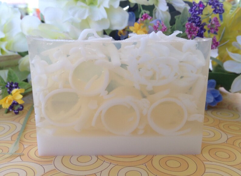 May include: A rectangular bar of soap with a clear, translucent top section filled with white, ring-shaped and shredded coconut pieces. The bottom section is opaque white. The background features flowers.