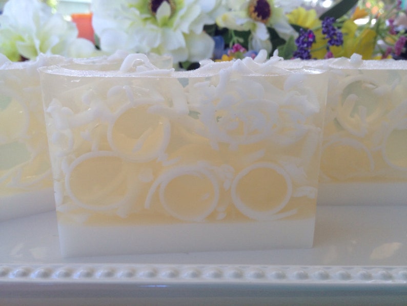 May include: Several bars of handmade soap with a layered design. The bottom layer is white, topped with a pale yellow layer, and a clear top layer with white decorative swirls. The soap is displayed on a white tray with a floral background.