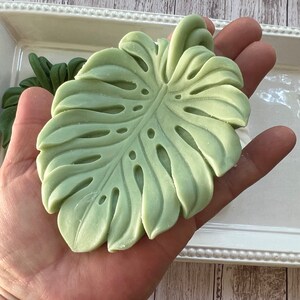 May include: A green, detailed, sculpted monstera leaf, held in a hand, against a white background.