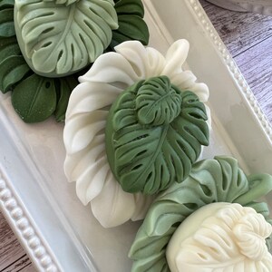 May include: Three decorative soap bars shaped like tropical leaves. The soaps are white and green and are arranged on a white tray with a beaded border.