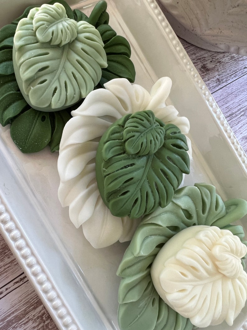 May include: Three decorative soap bars shaped like tropical leaves. The soaps are white and green and are arranged on a white tray with a beaded border.