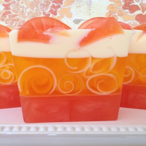 May include: Three bars of handmade soap with a layered design. The top layer is white, the middle layer is orange with white swirls, and the bottom layer is a bright red.