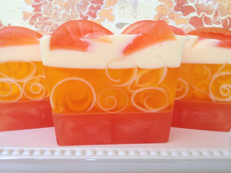 May include: Three bars of handmade soap with a layered design. The top layer is white, the middle layer is orange with white swirls, and the bottom layer is a bright red.