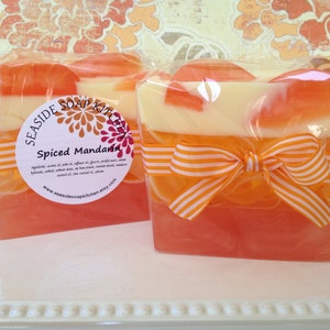 May include: Two bars of handmade soap wrapped in cellophane with orange and white striped ribbon. The soap is layered with orange, white, and a darker orange color. The label reads "Seashore Soap Kitchen" and "Spiced Mandarin".