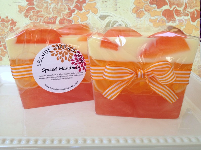 May include: Two bars of handmade soap wrapped in cellophane with orange and white striped ribbon. The soap is layered with orange, white, and a darker orange color. The label reads "Seashore Soap Kitchen" and "Spiced Mandarin".