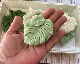 May include: A hand holding a green leaf-shaped soap bar. The soap is in the shape of a monstera leaf. There are other leaf-shaped soaps in the background.