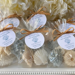 May include: Clear plastic favor bags containing assorted seashells and sea urchins, each tied with a gold ribbon and a round white tag printed with "SEASHELLS and wedding bells", "COURTNEY'S", "Bridal Shower", and "April 12, 2026" in blue text.