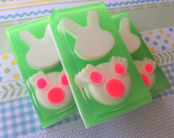 May include: Three green and white bunny-shaped soaps with pink accents. The soaps are made with a clear, glittery base and have a white bunny silhouette with pink paw prints.