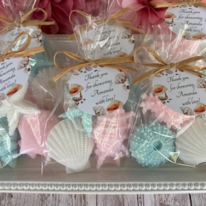 May include: A collection of seashell-shaped soaps in pastel colors, packaged in clear cellophane bags and tied with raffia bows. Each bag has a tag that reads "Thank you for showering Amanda with love!" The soaps are arranged on a white tray.