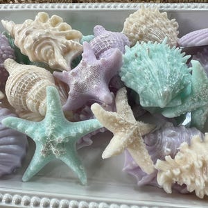 May include: A collection of assorted seashell and starfish shaped soaps in pastel shades of lavender, mint green, and cream. The items are arranged together in a rectangular white ceramic tray with a beaded edge detail along the rim.