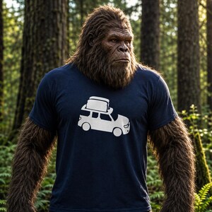 May include: A navy blue t-shirt featuring a white graphic of a car with a rooftop cargo carrier. The image shows a large, hairy creature, possibly Bigfoot, wearing the shirt while standing in a forest setting.