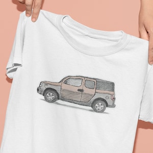 May include: White t-shirt featuring a sketch-style illustration of a brown and gray SUV. The vehicle has four wheels and a boxy design. The shirt is held up by a person's hands against a light pink background.