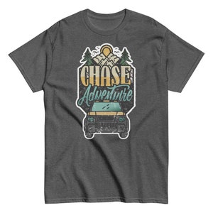 Può includere: Una t-shirt grigio scuro melange con una grafica in stile vintage di un'auto che guida attraverso un paesaggio montano. La grafica include il testo "CHASE Adventure" in un font usurato.